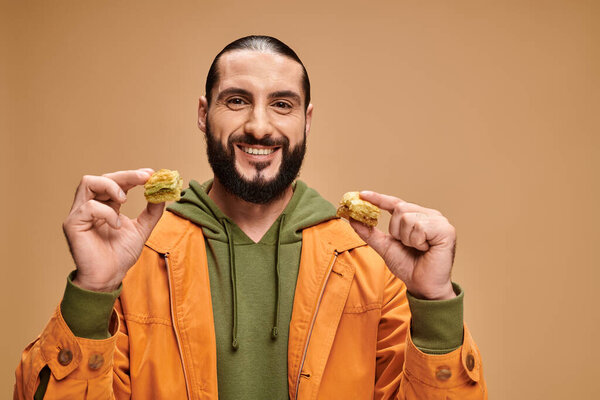 happy bearded man holding two different kinds of baklava on beige backdrop, turkish delights