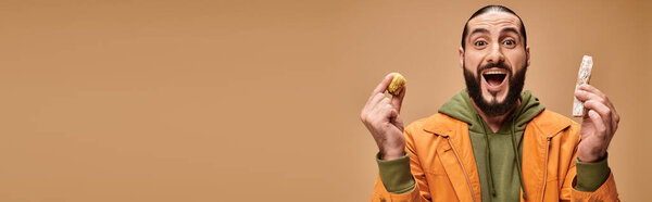 excited bearded man holding baklava and cevizli sucuk on beige backdrop, turkish delights banner
