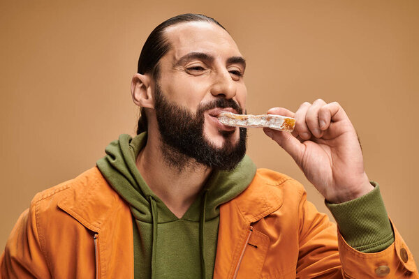 cheerful and bearded arabic man eating delicious cevizli sucuk on beige backdrop, turkish delights