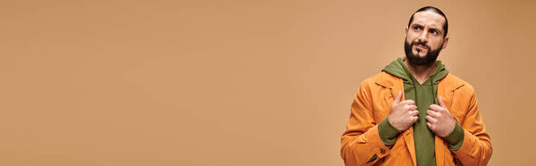 pensive middle eastern man with beard standing in casual attire on beige backdrop, banner