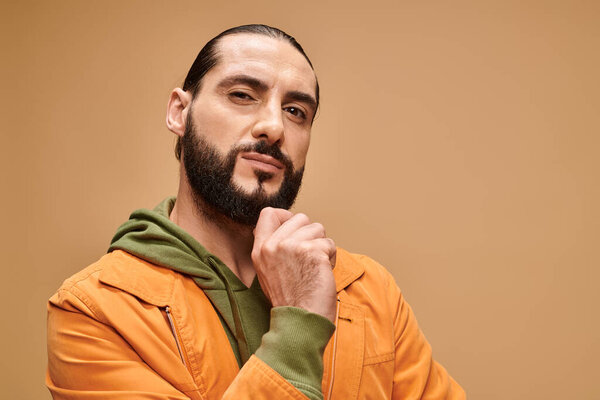 pensive middle eastern man touching beard and standing in casual attire on beige background