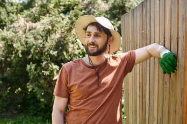 handsome and bearded farmer in sun hat standing near fence in countryside, rural lifestyle clipart