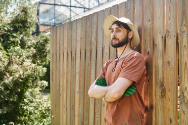 handsome and bearded farmer in sun hat standing with crossed arms near fence in countryside clipart