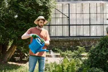Yakışıklı ve sakallı bahçıvan. Güneş şapkalı. Kırsaldaki seranın yakınındaki bitkileri suluyor.