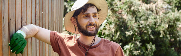 handsome and bearded farmer in sun hat standing near fence in countryside, rural lifestyle banner
