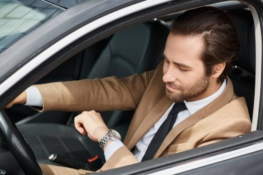 handsome bearded businessman in formal wear looking at wristwatch while driving car on street clipart