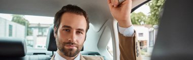 successful and handsome businessman in beige elegant suit looking at camera in luxury car, banner clipart