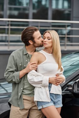 Yakışıklı adam, şehir sokağında modern arabanın yanında baştan çıkarıcı, şık, sarışın kadını kucaklıyor ve öpüyor.