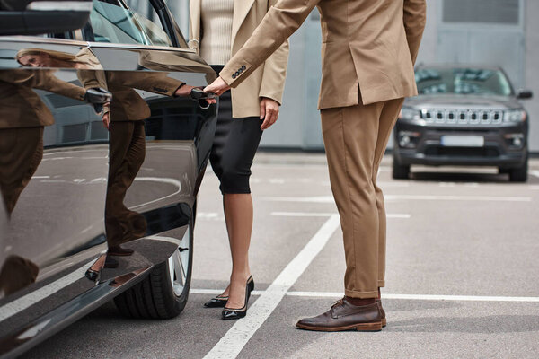 cropped view of man in stylish formal wear opening rear door of luxury car near businesswoman