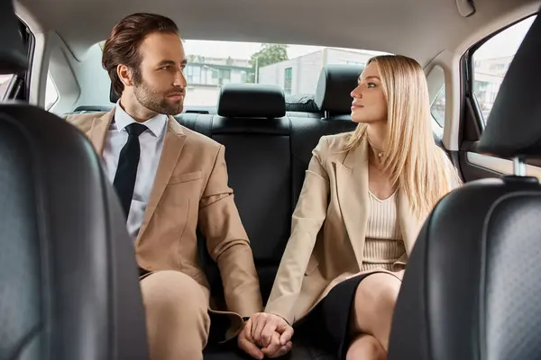 elegant business couple in formal wear holding hands and looking mat each other in car, attraction
