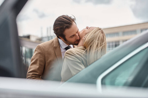 elegant businessman in formal wear embracing and kissing blonde woman near car on urban street