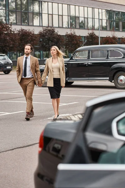 stock image elegant business couple in formal wear holding hands while walking along car parking, love affair