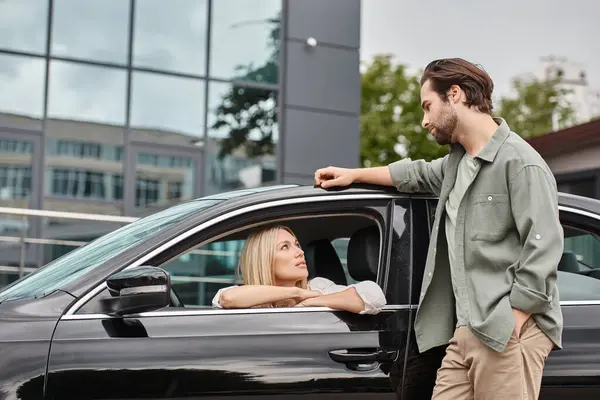 stock image handsome and stylish man standing near attractive blonde woman sitting in modern car, affection