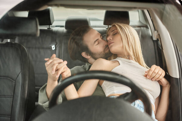 seductive and stylish couple embracing with closed eyes on drivers seat in modern car, love affair
