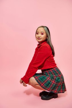 full length of young asian woman with heart shaped eye makeup and plaid skirt sitting on pink clipart