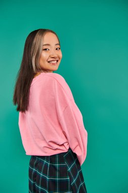 portrait of cheerful asian woman in pink sweatshirt and plaid skirt standing on blue backdrop clipart