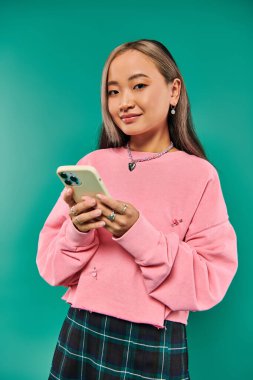 Pembe kazaklı mutlu Asyalı kadının portresi turkuaz arka planda akıllı telefon kullanıyor.