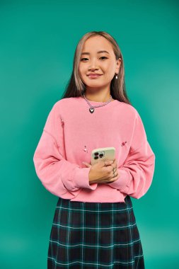Pembe kazaklı ve ekose etekli mutlu Asyalı kadının portresi turkuazda akıllı telefon kullanıyor.