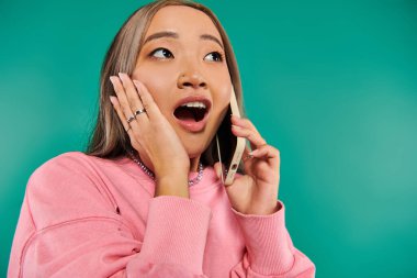 Pembe kazaklı genç Asyalı kadının portresi turkuaz arka planda akıllı telefondan konuşuyor.