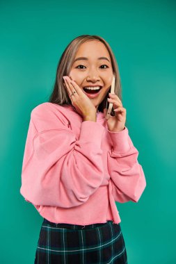Pembe sweatshirt 'lü, turkuaz arka planda akıllı telefondan konuşan Asyalı genç kadının portresi.