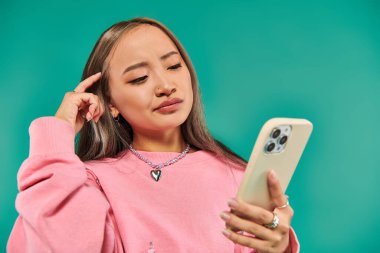 Mavi üzerine akıllı telefon kullanarak pembe kazak ve ekose etek giyen düşünceli Asyalı genç kadının portresi.