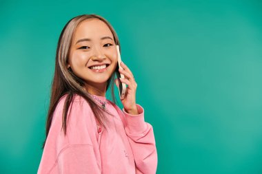 Heyecanlı pembe kazaklı genç Asyalı kadın turkuaz arka planda akıllı telefondan konuşuyor.