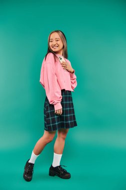 happy young asian woman in sweatshirt and plaid skirt holding smartphone on turquoise backdrop clipart