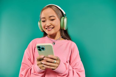 Kablosuz kulaklıklı neşeli Asyalı kız müzik dinliyor ve turkuaz arka planda akıllı telefon kullanıyor.