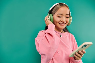 Kablosuz kulaklıklı neşeli Asyalı kız müzik dinliyor ve turkuazda akıllı telefon kullanıyor.