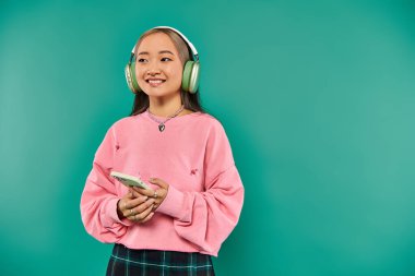 Kablosuz kulaklıklı neşeli Asyalı kadın müzik dinliyor ve turkuaz fon üzerinde akıllı telefon kullanıyor.