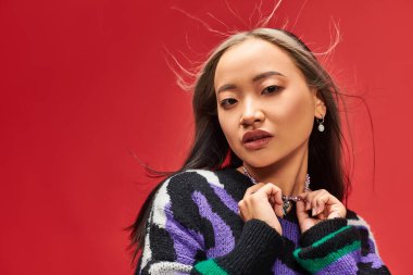 alluring young asian woman in vibrant sweater with animal print touching necklace on red backdrop clipart