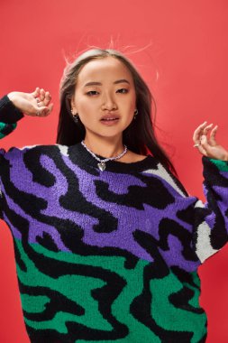 young asian girl in sweater with animal print with heart shaped necklace posing on red backdrop clipart