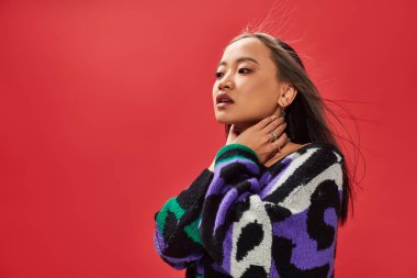 Kırmızı arka planda poz verirken hayvan desenli süveter giyen güzel Asyalı kadın boynuna dokunuyor.
