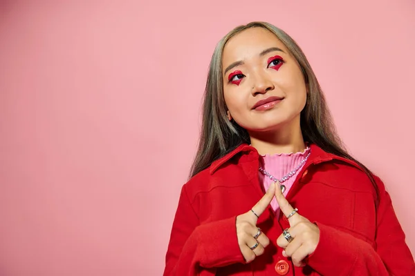 stock image pleased young asian woman with heart shaped eye makeup smiling and looking away on pink backdrop