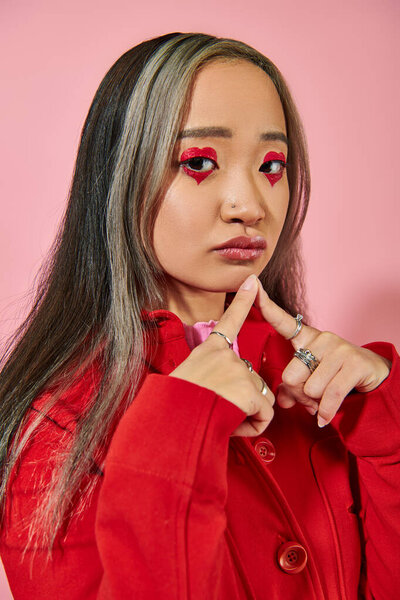 pensive young asian woman with heart shaped eye makeup and dyed hair looking at camera on pink