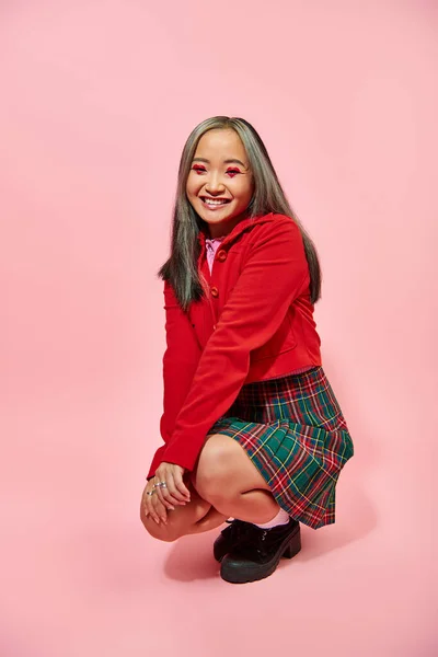 stock image full length of cheerful asian woman with heart shaped eye makeup and plaid skirt sitting on pink