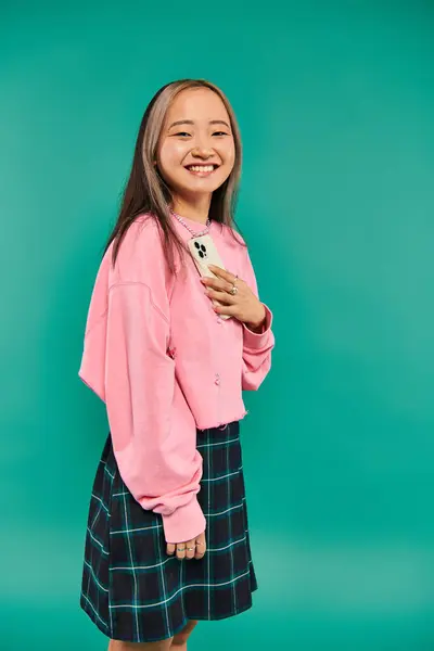 stock image portrait of positive young asian woman in pink sweatshirt and plaid skirt holding smartphone on blue