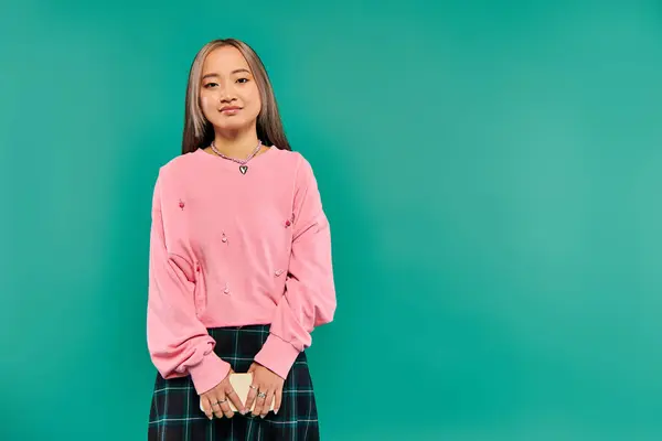 stock image beautiful young asian woman in sweatshirt and plaid skirt holding smartphone on turquoise backdrop