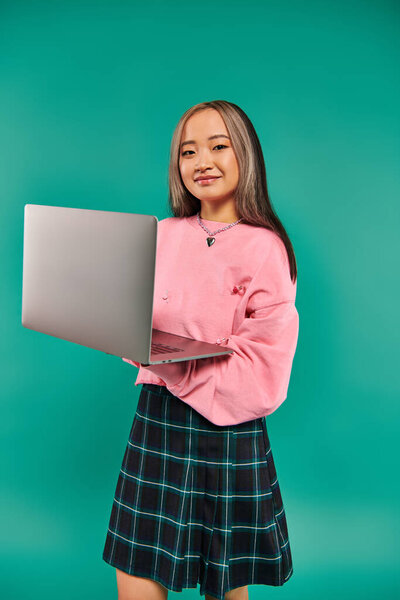 happy asian girl in pink sweatshirt using laptop while standing on turquoise backdrop, networking
