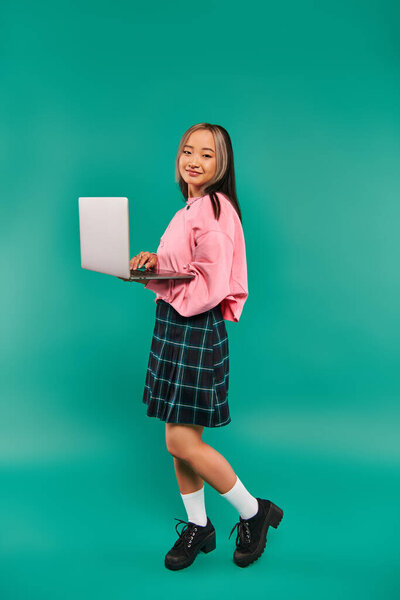 pleased asian girl in pink sweatshirt using laptop while standing on turquoise backdrop, networking
