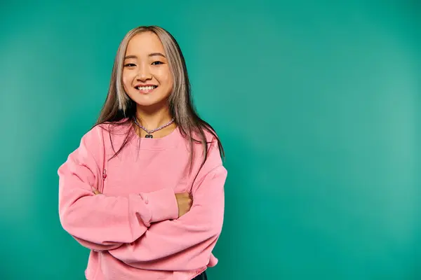 stock image portrait of pleased young asian girl in pink sweatshirt standing with crossed arms on turquoise