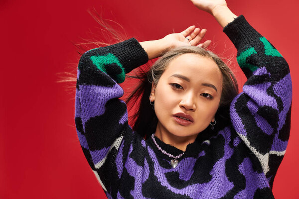 привлекательная молодая азиатка в ярком свитере с отпечатком животного, позирующая с поднятыми руками на красном