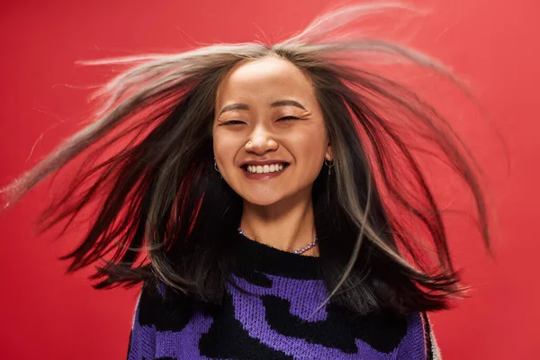 wind in hair of joyful young asian woman in sweater with animal print posing on red background