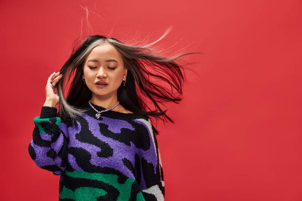wind in hair of pretty young asian girl in sweater with animal print posing on red background