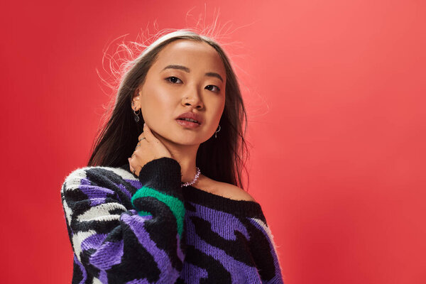 young pretty asian woman in sweater with animal print touching neck while posing on red backdrop