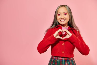 Valentines day, happy asian girl with red eye makeup showing heart with hands on pink background clipart