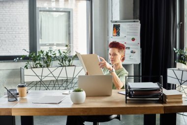 Ofiste kağıt bardağa koymak için laptopun ve kahvenin yakınındaki belgelerle çalışan şık biseksüel yönetici