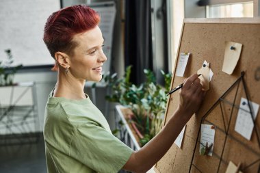 smiley bigender manager in casual attire writing on paper notes on corkboard in modern office clipart