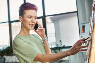 stylish queer person talking on mobile phone and looking at paper notes on corkboard in office clipart