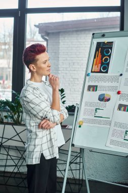 thoughtful bigender person looking at flip chart with business analytics in while working in office clipart